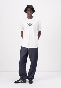 Witte katoenen t-shirt met zwart Adidas-logo, donkerblauwe spijkerbroek en zwarte bucket hat. Draag het met zwart-witte sneakers.