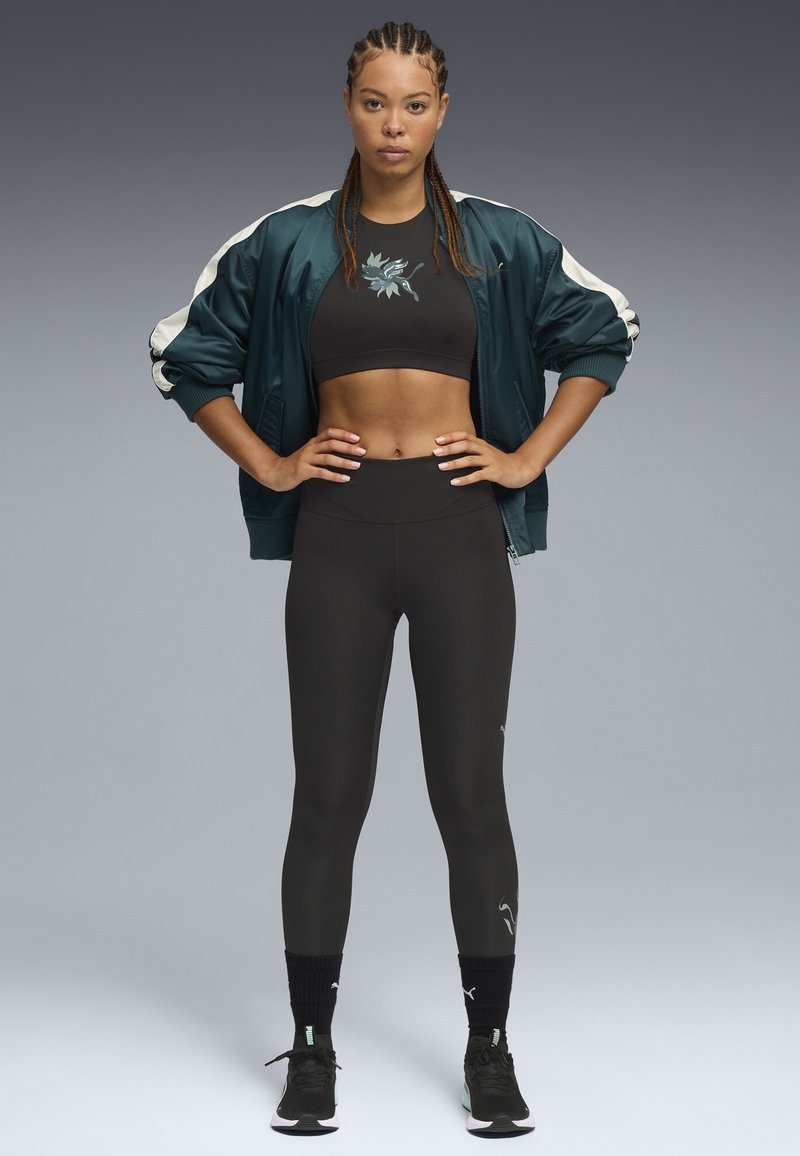 Sujetador deportivo negro, chaqueta bomber de satén verde oscuro con rayas crema, leggings negros de cintura alta y zapatillas deportivas negras con detalles en turquoise.