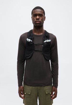 Jonge man draagt een zwart hydratatievest met twee waterflessen over een donker shirt met lange mouwen en groene cargoshorts, staand frontaal.