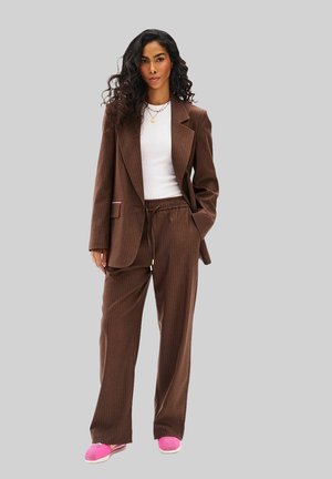 Kobieta z kręconymi włosami ubrana w brązowy garnitur w prążki, biały top, warstwowe naszyjniki i różowe trampki, stojąca z jedną ręką w kieszeni.