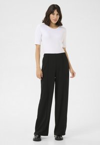 Weißes Kurzarmoberteil kombiniert mit hochgeschnittenen, weiten schwarzen Hosen. Glattes Material mit taillierter Passform und mittleren Falten. Schwarze Schuhe.