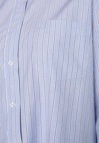 Camisa botão em azul claro com riscas verticais estreitas em tons mais escuros, apresentando um bolso no lado esquerdo do peito e uma textura de tecido suave.