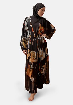 Kvinde med sort hijab bærer en lang sort blomsterdragt med orange og beige blomster, står barfodet mod en ensfarvet baggrund.