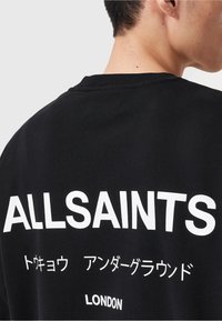 Person wearing black shirt with white text "ALLSAINTS," Japanese characters, and "LONDON" on the back, shown from shoulder to neck.