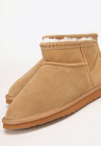 Tan suede ankle boots with white fur lining and brown rubber soles, designed for warmth and indoor or outdoor comfort.