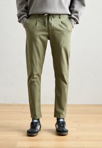 Pantaloni verzi măslinii cu talie cu șnur, design pliat și buzunare laterale, asortați cu pantofi negri cu accente albe.