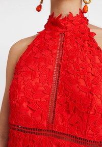 Robe rouge à encolure halter avec broderie en dentelle florale, détail en trou de serrure à l'avant et boucles d'oreilles pendantes oranges assorties, portée par une personne avec les épaules nues.