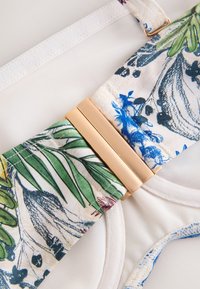 Maillot de bain en tissu mesh blanc avec des bretelles fleuries ornées d'imprimés botaniques verts, bleus et noirs, reliées par une boucle rectangulaire beige.