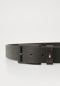 Ceinture en cuir marron avec une texture lisse, dotée d'une boucle en métal noir et d'un petit logo en rouge, blanc et bleu.