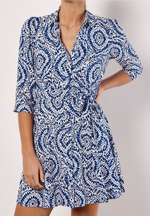 Femme portant une robe blanche avec des motifs tie-dye circulaires bleus et des manches trois-quarts, posant avec une main sur la hanche.