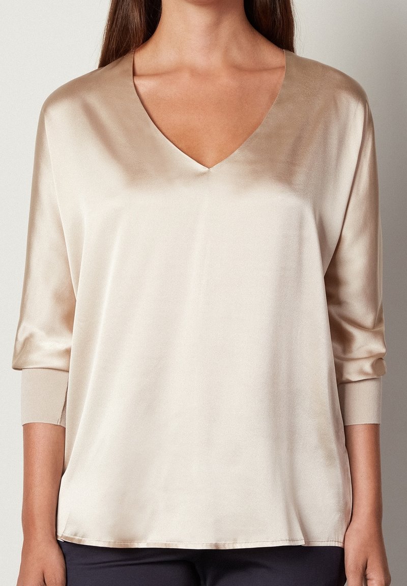 Femme portant un chemisier en satin beige avec des manches trois quarts et un col en V, associé à un pantalon foncé, montrée du cou aux hanches.