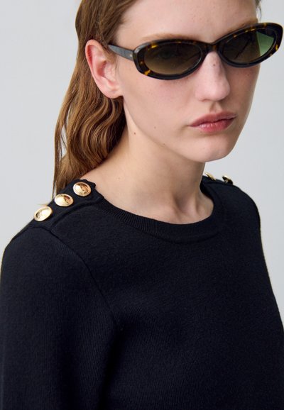 Pull nera con finitura texturizzata, caratterizzato da bottoni dorati sulle spalle. Il modello indossa occhiali da sole scuri e ha i capelli sciolti.