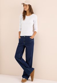 Weißes Langarm-T-Shirt, dunkelblaue Jeans mit weitem Bein, beiger Hut und hellbraune Sneakers; lässiger, entspannter Schnitt mit Taschen und sichtbaren Nähten.