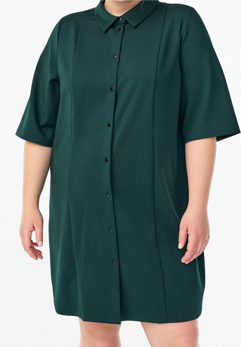 Robe-chemise vert foncé à manches courtes avec col et boutons noirs portée par une personne debout devant un fond blanc.