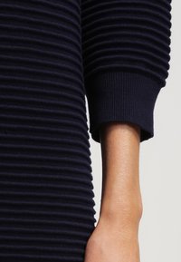 Bras portant un pull bleu marine côtelé à manches longues avec un poignet serré, sur un fond clair uni.