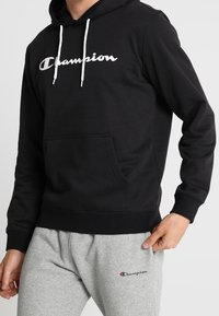 Felpa nera con logo bianco "Champion", con coulisse e tasca a marsupio; indossata con pantaloni della tuta grigi con un piccolo logo.