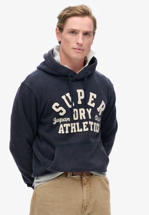 Marineblauwe hoodie met beige tekst, met "SUPER DRY ATHLETICS" en een kangoeroezak. Gelaagd over een lichtgrijs shirt.