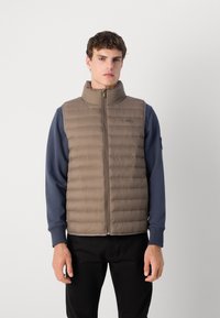 Bruine gewatteerde vest met hoge kraag, frontzipper en zijzakken. Draagt over een blauwe shirt met lange mouwen, met een soepele textuur en een gequilt ontwerp.