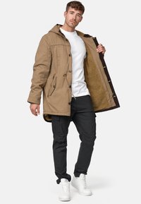 Veste parka beige avec capuche, dotée d'une fermeture à boutons, d'une doublure en polaire et de deux poches avant. Associée à un pantalon cargo noir et des baskets blanches.