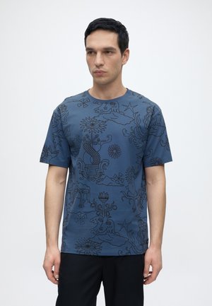 Homme aux cheveux noirs courts portant un t-shirt bleu avec un dessin abstrait en lignes noires et un pantalon noir, debout devant un fond blanc uni.