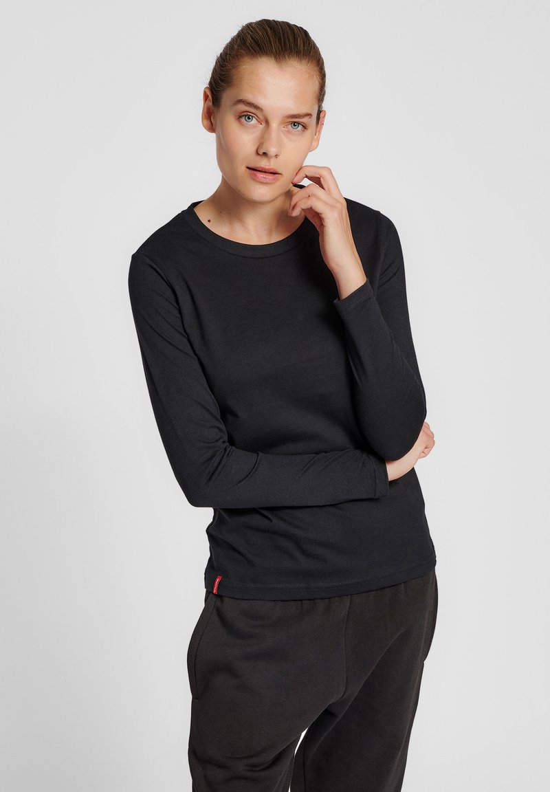 Frau mit heller Haut und zurückgebundenem Haar, die ein schwarzes Langarmshirt und dunkle Hose trägt und mit einer Hand nahe ihrem Gesicht posiert.