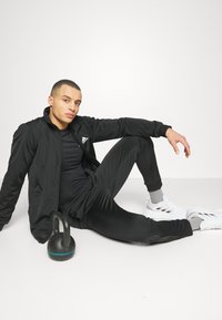 Black athletic tracksuit with a zip jacket, fitted top, and tapered pants. Black kettlebell beside the subject. White sneakers with gray accents.