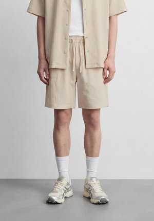 Osoba oblečená v béžových štruktúrovaných krátkych nohaviciach a zodpovedajúcej košeli s krátkym rukávom cez biele tričko, s bielymi členkovými ponožkami a béžovými teniskami stojaca na sivej podlahe.