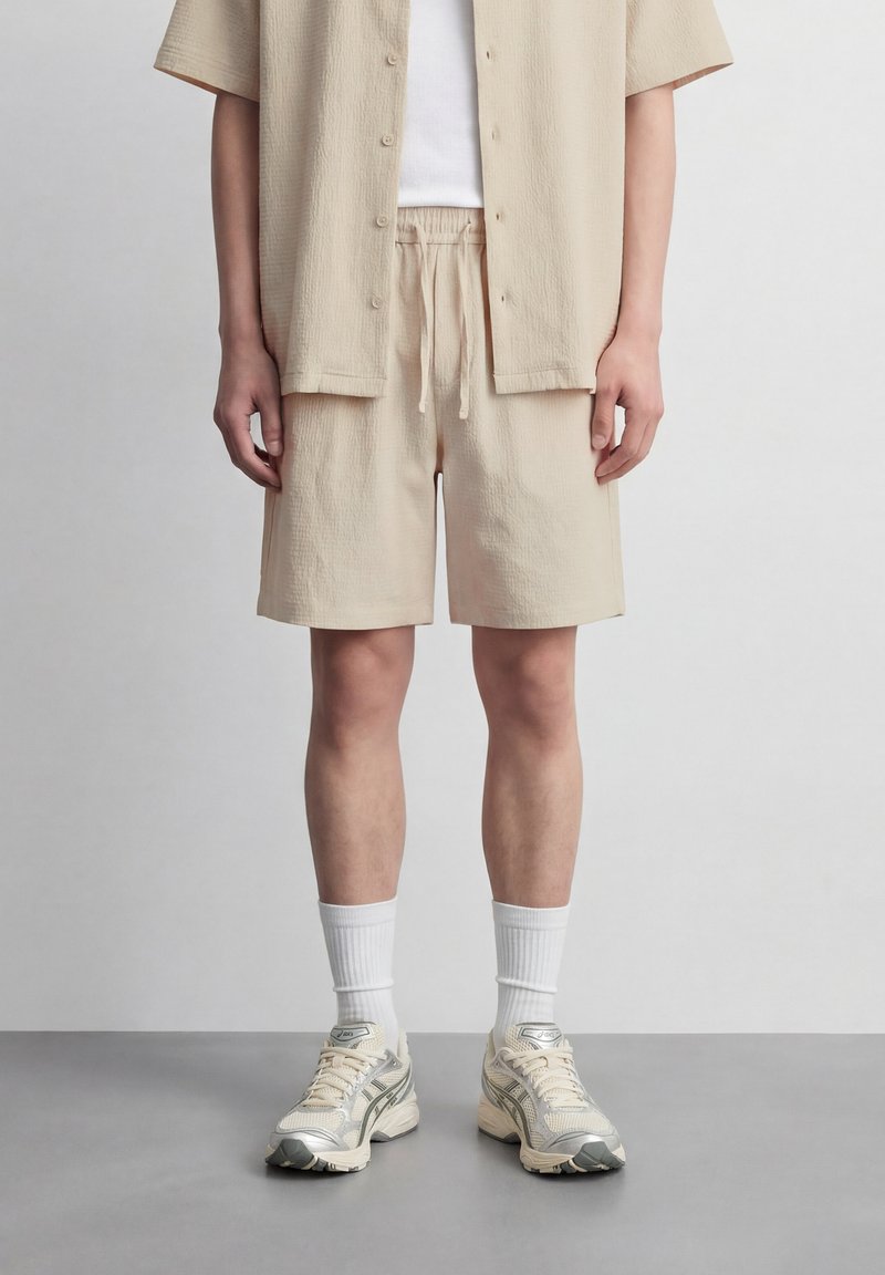 Άτομο που φοράει μπεζ σορτς με υφή και ταιριαστό κοντομάνικο πουκάμισο πάνω από λευκό τοπ, λευκές κάλτσες μέσης και μπεζ αθλητικά παπούτσια, στέκεται σε γκρι δάπεδο.