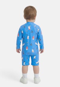 Malček z svetlo rjavimi lasmi, oblečen v modro dolgoročno oblačilo z risanimi liki in belimi supergami, obrnjen stran.