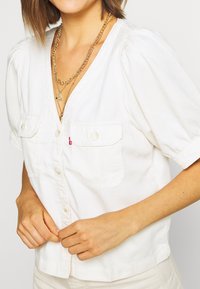 Chemise blanche en coton avec boutons, manches courtes bouffantes, deux poches poitrine et un petit logo rouge sur la poche gauche.