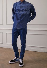 Denim-paita, jossa on edessä logo-printti, yhdistettynä sinisiin farkkuihin ja mustiin lenkkareihin, joissa on värikkäitä yksityiskohtia ja valkoiset nauhat.