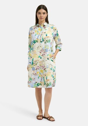 Frau trägt ein helles, knielanges Hemdblusenkleid mit buntem Blumen- und Gebäudemuster, steht mit einer Hand in der Tasche und braunen Sandalen.