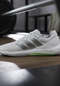 Chaussure de sport blanche avec des rayures grises et un détail vert sur la semelle, placée sur le sol de la salle de sport près d'une barre d'haltères dans un cadre d’entraînement.