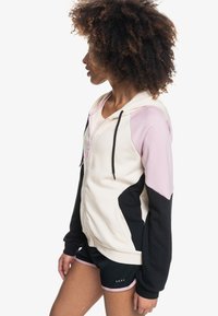 Sudadera con capucha en colores crema, rosa y negro; cuenta con un cierre frontal, bloques de color contrastantes y puños elásticos. La tela parece suave.