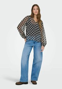 Jeune femme portant un chemisier transparent à pois noirs avec des manches longues, un jean large bleu clair et des chaussures marron, se tenant détendue.