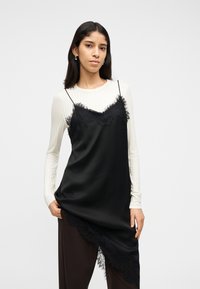 Γυναίκα με μακριά σκούρα μαλλιά φοράει λευκό μακρυμάνικο τοπ κάτω από μαύρο φόρεμα με δαντελένιο τελείωμα, στέκεται μπροστά σε απλό φωτεινό φόντο.