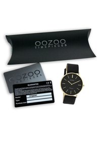 Montre noire avec des accents dorés et un cadran rond, accompagnée d'une boîte d'emballage noire et de deux cartes de garantie.