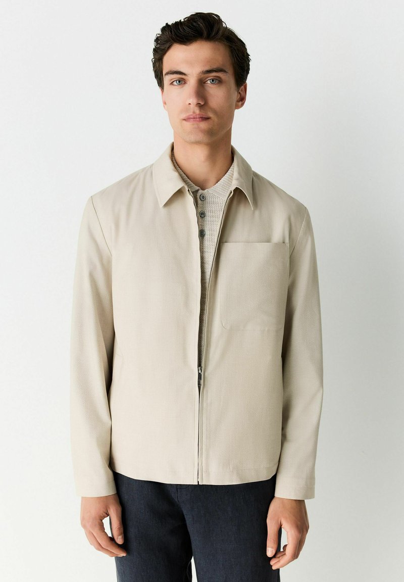 Chaqueta beige con cierre frontal, cuello puntiagudo y un bolsillo en el pecho. Lleva puesta sobre una camisa gris claro texturizada, combinada con pantalones oscuros.