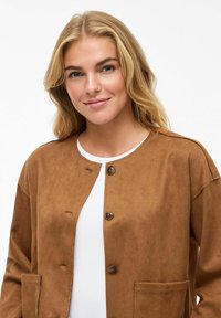 Veste en daim marron avec un col arrondi, fermeture à boutons sur le devant et deux poches latérales. Texture lisse et conception à coupe décontractée.
