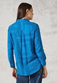 Camisa azul de manga larga con patrones cuadrados negros, fabricada en un tejido ligero, vista desde atrás, combinada con jeans oscuros, mostrando un ajuste suelto.