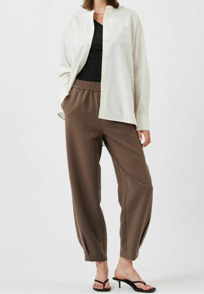 Personne portant un pantalon marron ample avec taille élastique, un haut noir, une chemise ample écrue boutonnée et des sandales noires à talons.