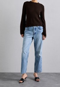 Brązowa teksturowana bluzka z długim rękawem i falowanym dołem, zestawiona z jasnoniebieskimi jeansami o prostych nogawkach oraz czarnymi sandałami na obcasie typu slide.