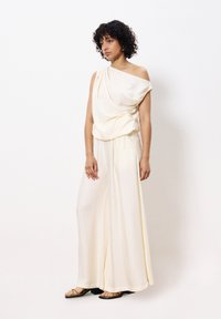Roomkleurige top met één schouder en gedrapeerd stof, gecombineerd met een broek met wijde pijpen. Lichtgewicht, textuurmateriaal. Platte sandalen.
