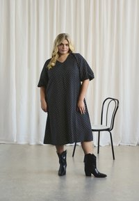 Robe noire à pois avec un décolleté en V et de courtes manches ballons. Une paire de bottes de cowboy noires complète la tenue. Fond minimaliste.