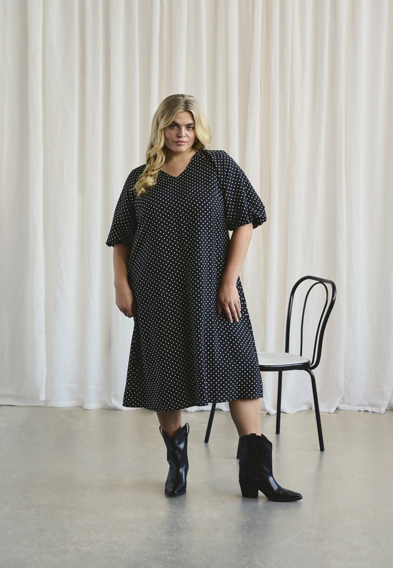 Robe noire à pois avec un décolleté en V et de courtes manches ballons. Une paire de bottes de cowboy noires complète la tenue. Fond minimaliste.
