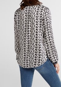 Svart och vit mönstrad långärmad skjorta med textdesign, rak nederkant, sidoklipp och mjukt tyg. Kombinerad med blå denimjeans.