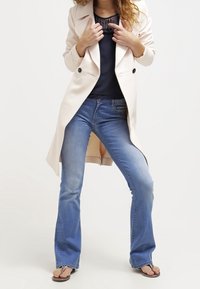 Cappotto lungo color beige chiaro con bottoni neri, indossato sopra un top blu navy e jeans a campana blu. Abbinato a ciabatte scure. Texture del tessuto morbida.