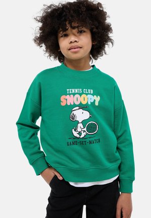 Enfant aux cheveux bouclés portant un sweat-shirt vert avec Snoopy tenant une raquette de tennis et le texte « Tennis Club Snoopy Game-Set-Match ».