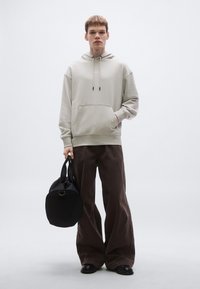 Helehall kapuutsiga dressipluus eesmise taskuga, kombineeritud lahtiste pruuni tooniga pükstega, hoides ümmargust musta kotti. Silmapaistev siledus, mugav lõige.