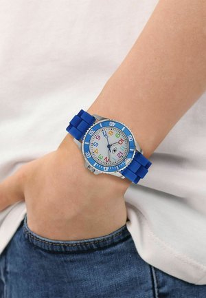 Wrist watch with blue rubber strap and colorful numbers on white dial worn on left wrist against white shirt and blue jeans.